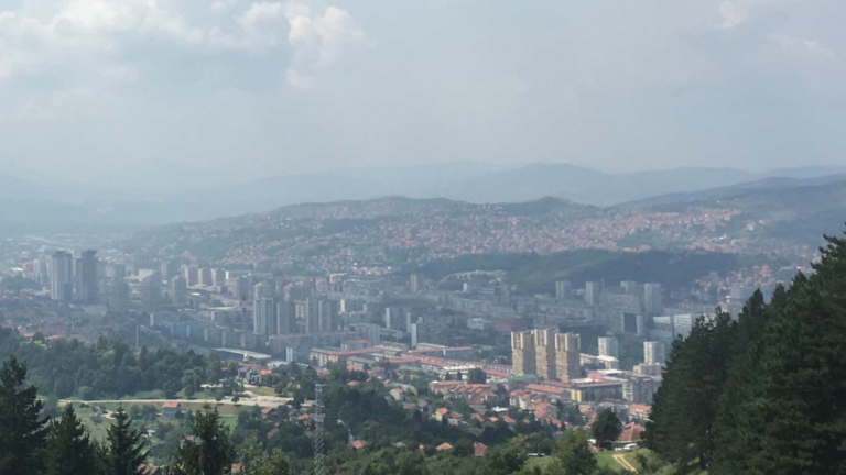 An aerial view of Sarajevo. We see trees in the foreground and orange-roofed buildings nestled amongst hills and mountains.